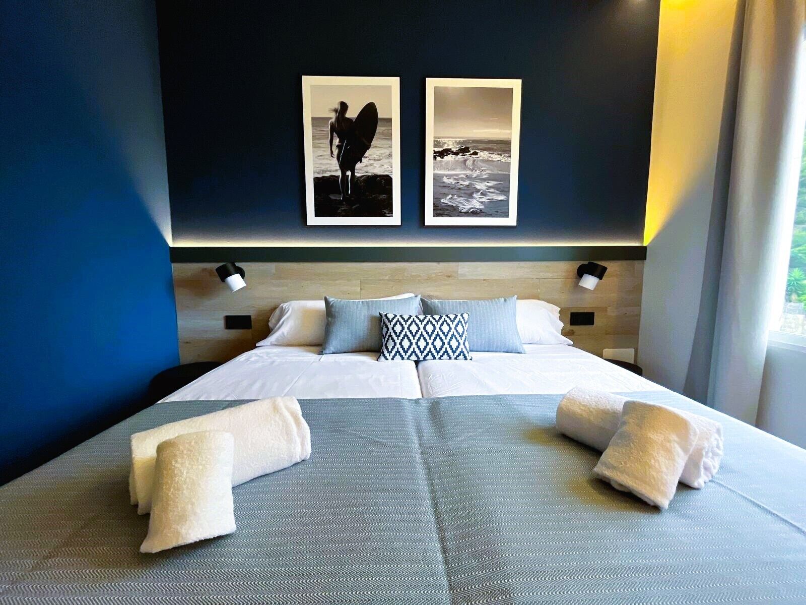 A modern hotel room with a large bed, two wall-mounted lamps, and two framed pictures depicting beach scenes.