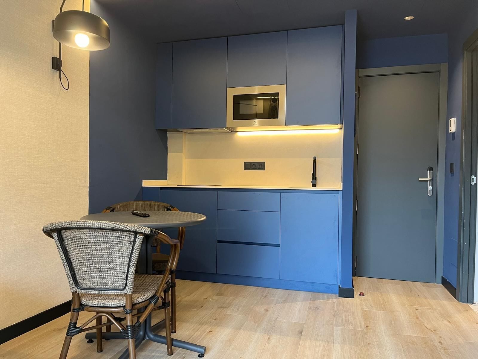 A compact kitchen with blue cabinets, a microwave, and a small dining area with a wicker chair and a round table.