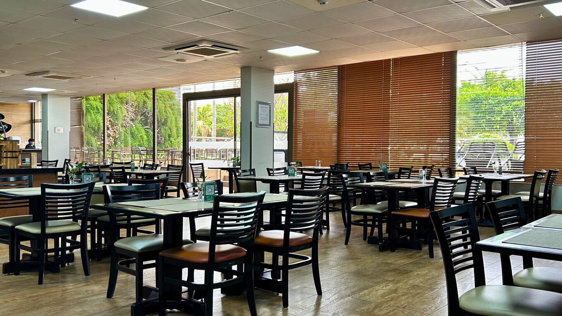 A spacious indoor dining area with multiple tables and chairs, large windows with blinds, and a view of greenery outside.