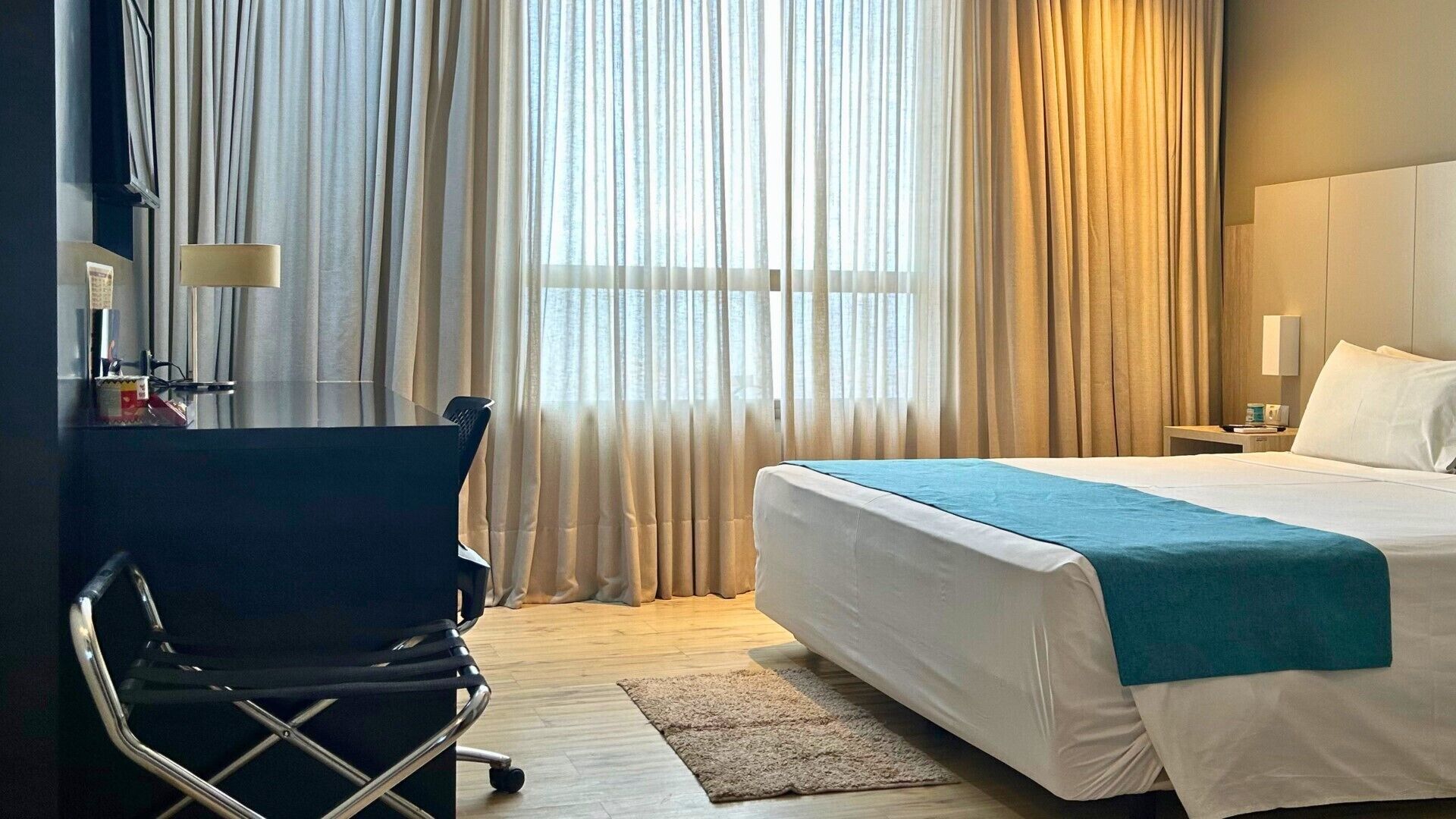 A hotel room with a bed, a desk with a chair, a nightstand, and a window with curtains.