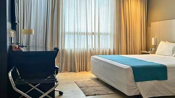 A hotel room with a bed, a desk with a chair, a nightstand, and a window with curtains.