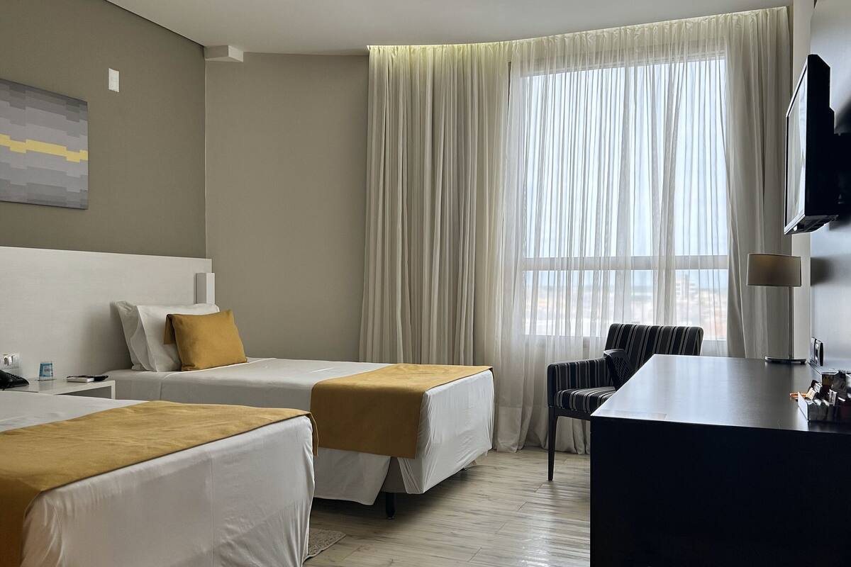 A hotel room with two beds, a desk, a chair, a TV, and a window with curtains.
