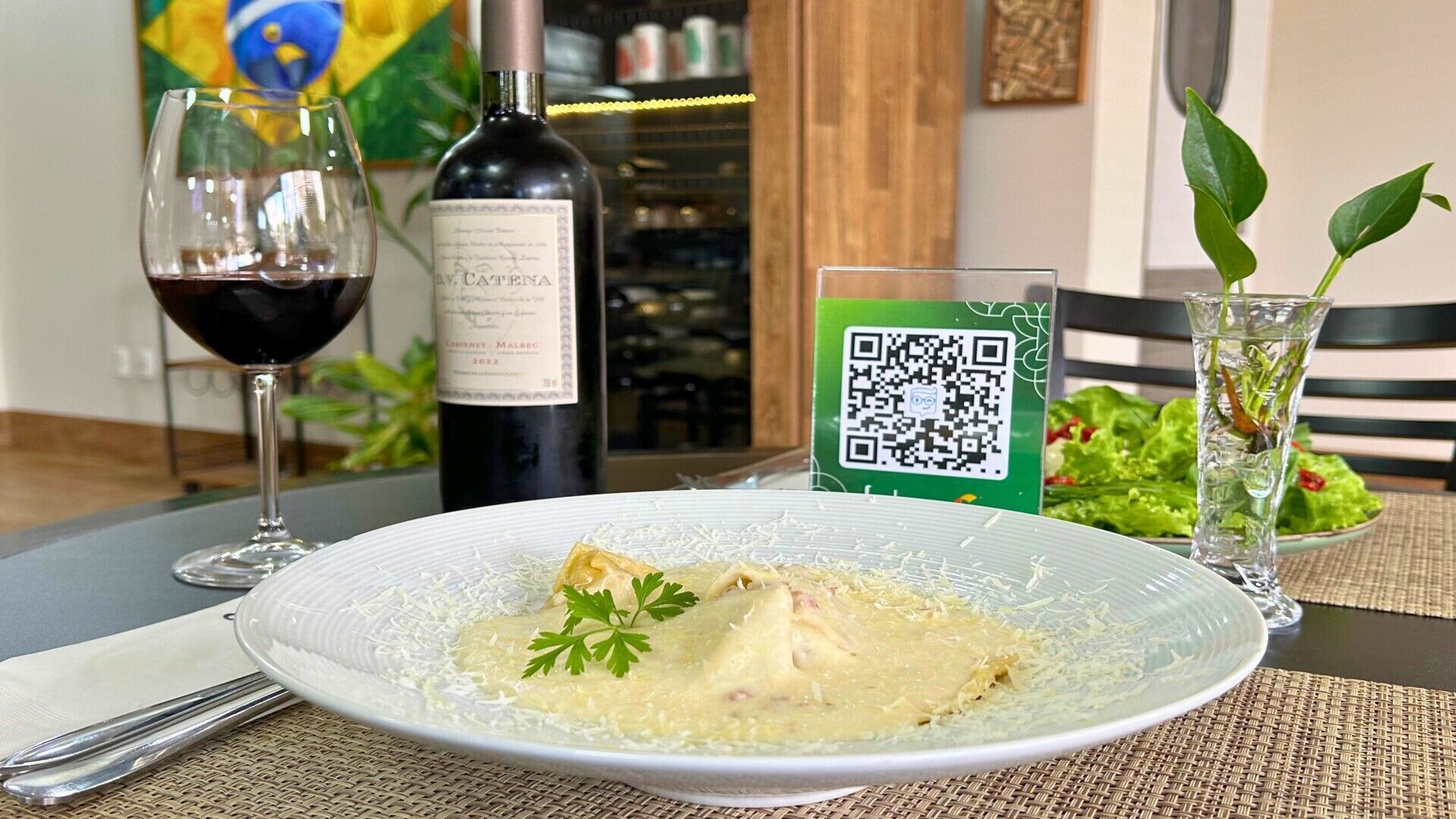 A dining table with a plate of food, a wine glass, a bottle of wine, a QR code sign, and a glass of water.