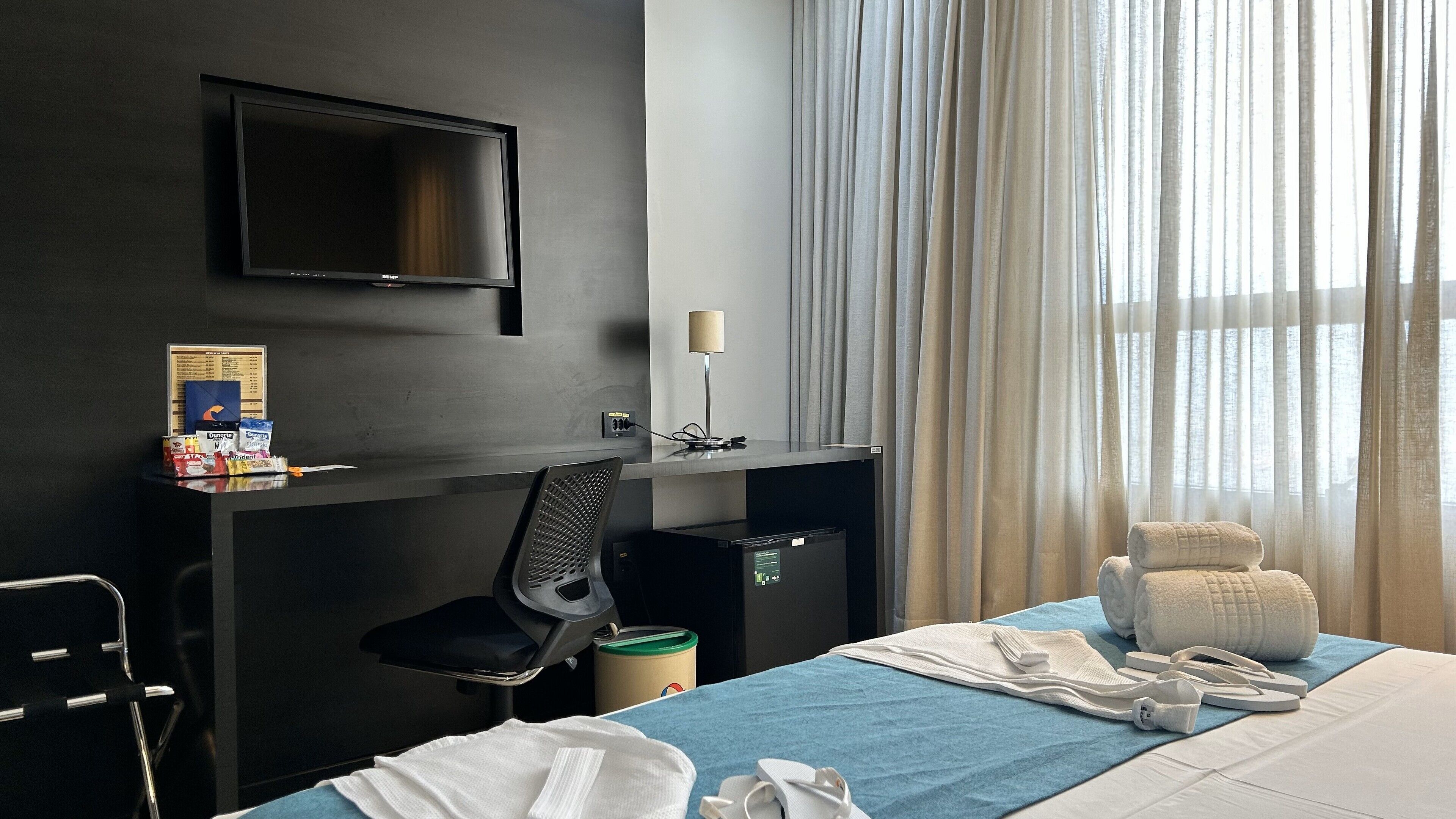 A hotel room with a bed, a desk with a computer, a chair, a TV mounted on the wall, and a window with sheer curtains.