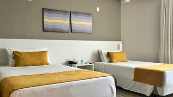 Quarto de hotel com duas camas, cada uma com roupa de cama branca e almofadas cor mostarda. Existem duas obras de arte fixadas na parede acima das camas, uma mesa de cabeceira com telefone e máquina de café, e uma janela com cortinas.