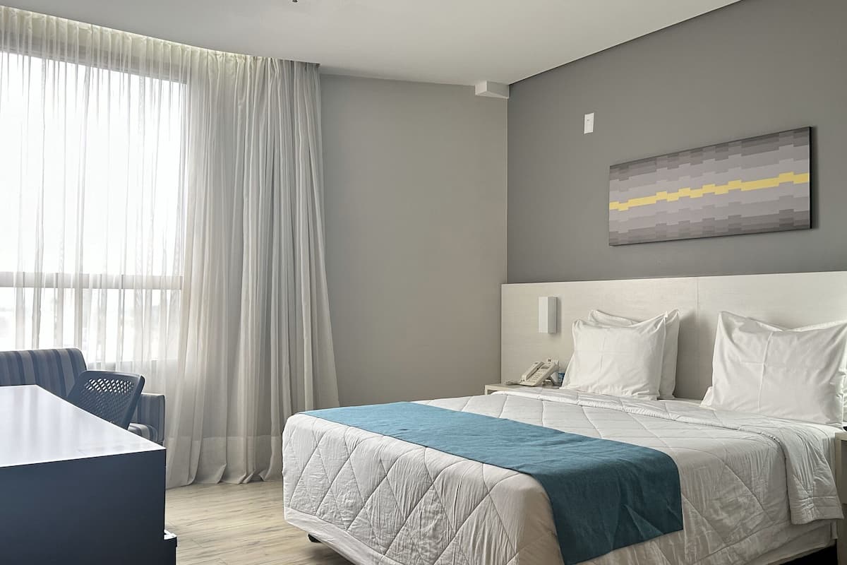 A hotel room with a bed, a desk, a chair, a bedside table with a phone, and a painting on the wall.