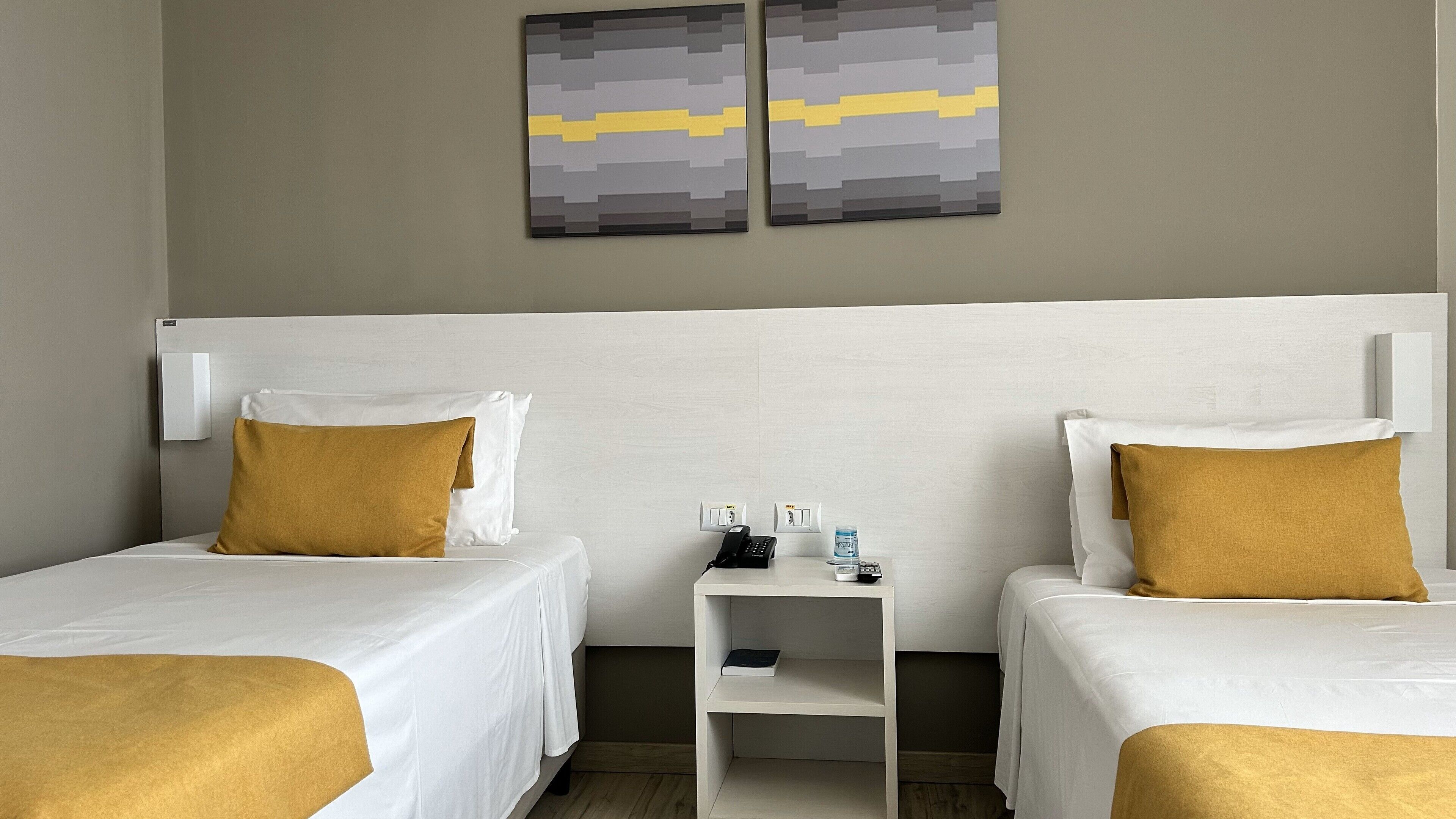 A hotel room with two beds, each with white bedding and a mustard-colored pillow. There is a small bedside table with a phone and a bottle of water. The walls are painted in a neutral color, and there are two abstract art pieces above the beds.