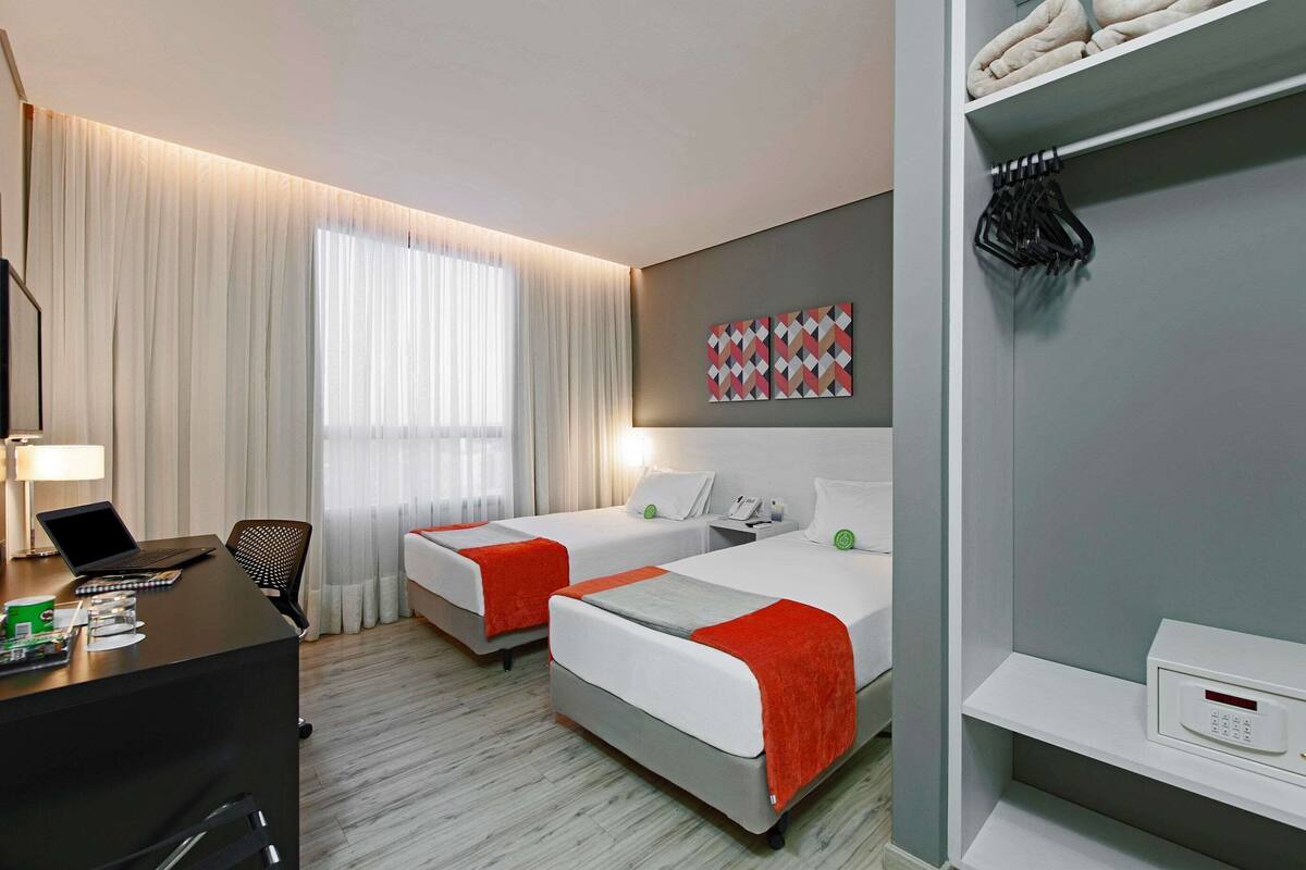 A hotel room with two beds, a desk with a laptop, a chair, a desk lamp, a window with curtains, a wall-mounted artwork, and a shelf with towels and a control panel.