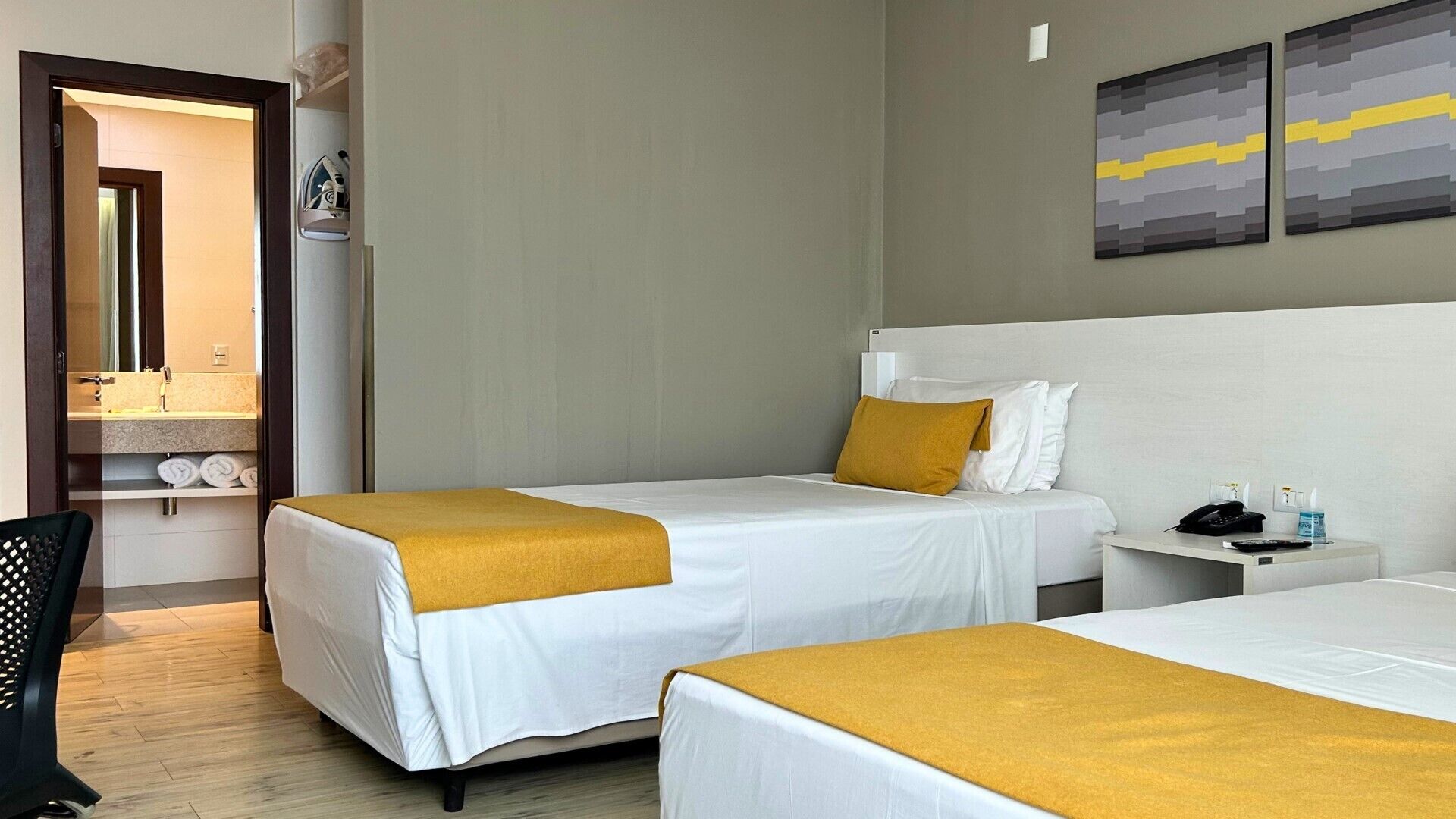 Habitación de hotel con dos camas, una mesita de noche con un teléfono y una botella, y un baño visible a través de una puerta abierta.