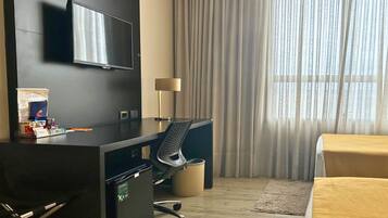 A hotel room with a desk, chair, TV, lamp, and a bed.