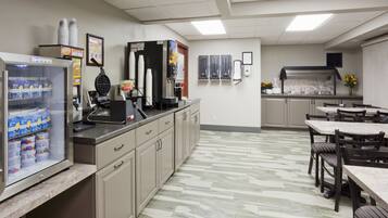 A clean, well-lit cafeteria with a self-service beverage station, a refrigerator stocked with beverages, and a tiled floor.