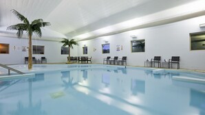 An indoor swimming pool with clear water, a palm tree, and several chairs along the poolside.