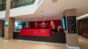 Un'area reception di un hotel con un banco rosso, un cartello che indica servizi come bar self-service, ristorante e centro business, e una scala che conduce a un piano superiore.