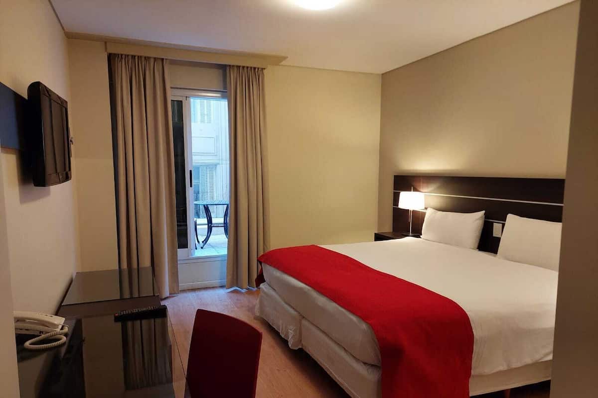 A hotel room with a bed, a desk, a chair, a TV, and a balcony with curtains.