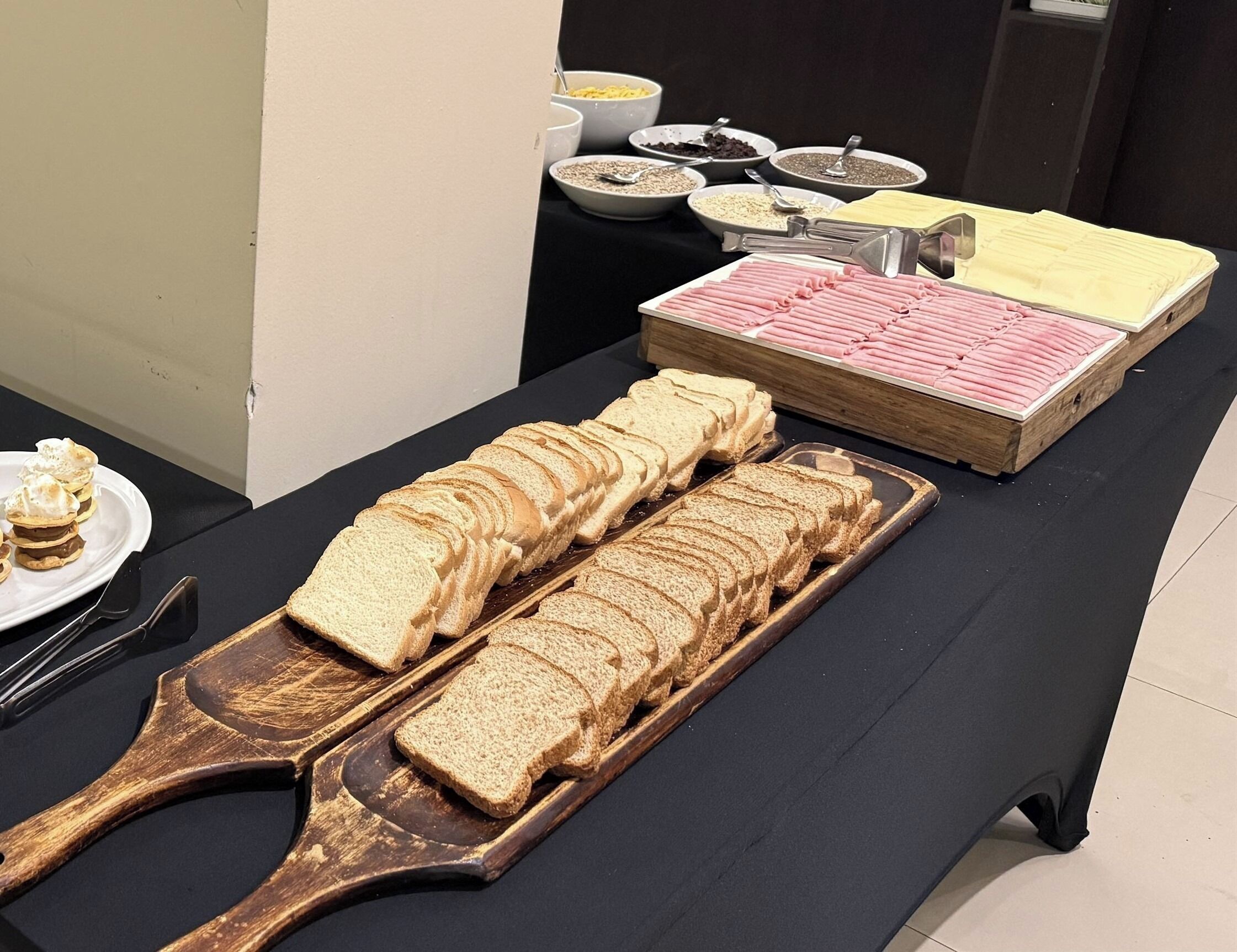 A buffet table with sliced bread, a wooden serving board, and various food containers.