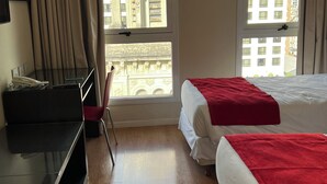 Una camera d'hotel con due letti, una scrivania, una sedia e ampie finestre che offrono vista sugli edifici.