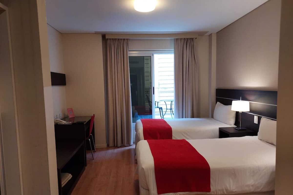 A hotel room with two beds, a desk, a chair, and a view of the outside.
