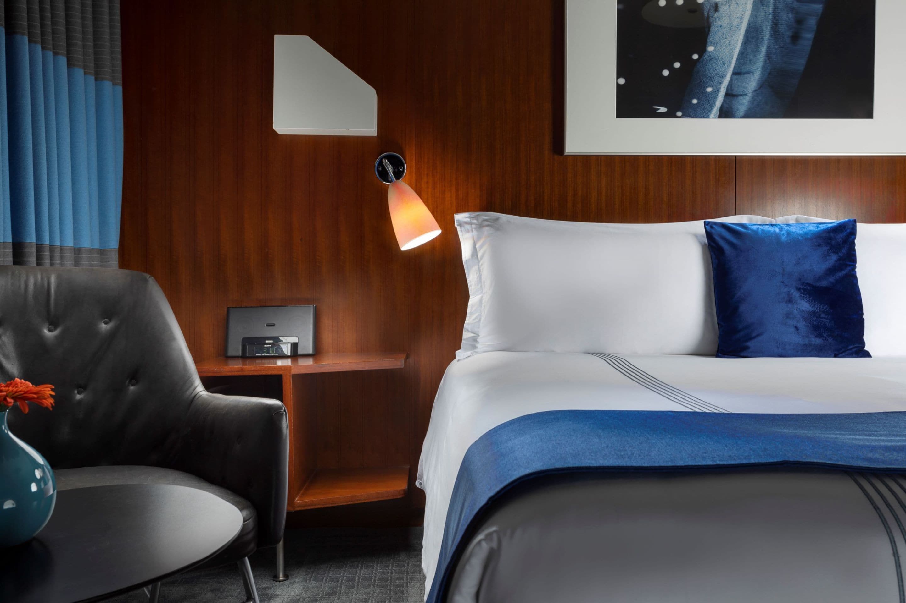 A hotel room with a bed, a nightstand, a desk, a chair, and a wall-mounted lamp.