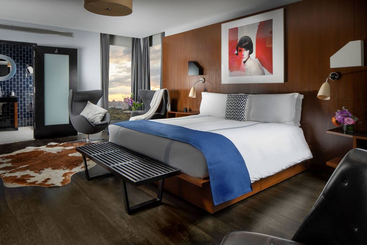 A modern hotel room with a large bed, a sitting area with a chair and a small table, a view of the city through large windows, and a wooden accent wall.