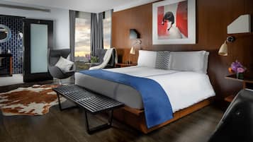A modern hotel room with a large bed, a sitting area with a chair and a small table, a view of the city through large windows, and a wooden accent wall.