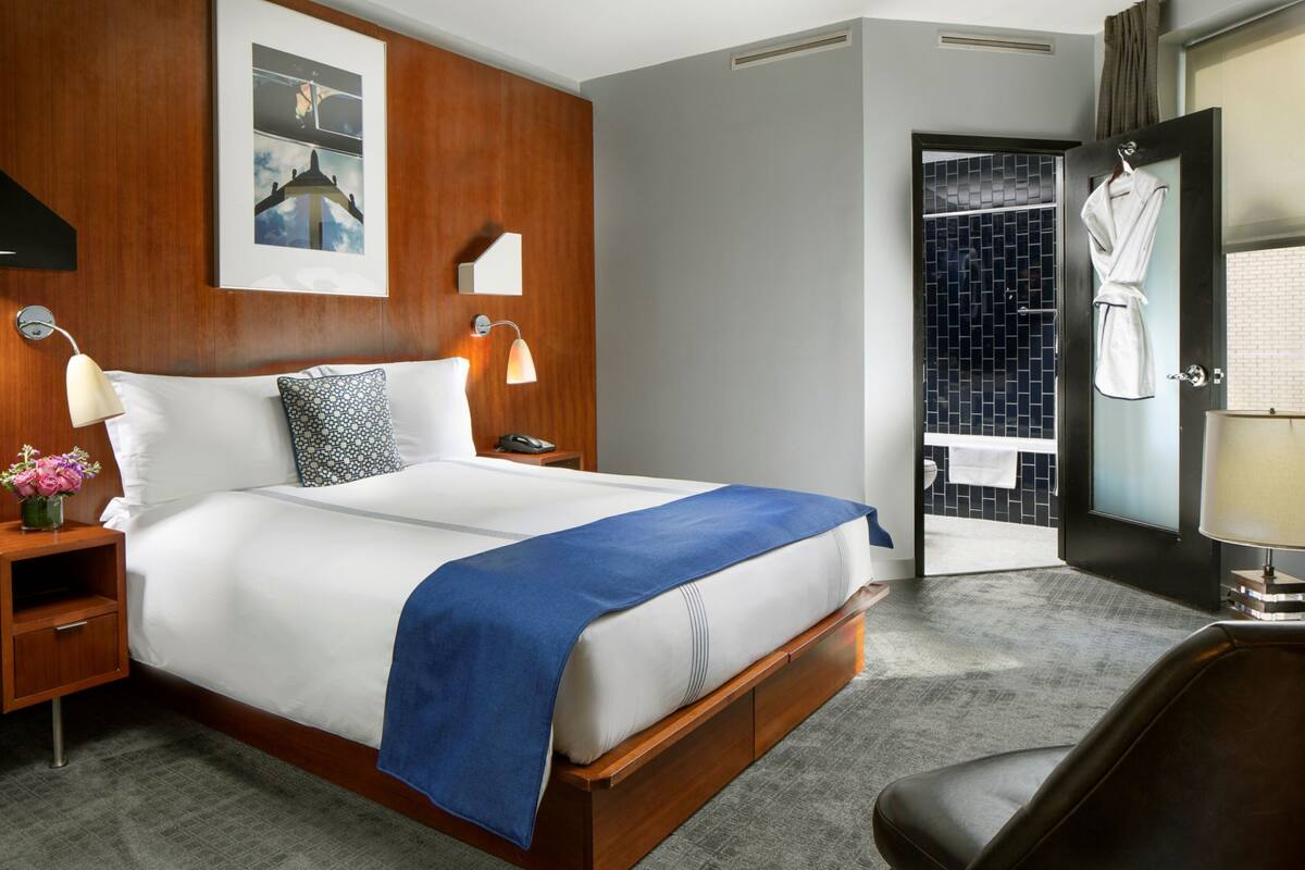A hotel room with a large bed, a wooden headboard, a nightstand with a lamp, a bathroom with a shower, and a robe hanging on the door.