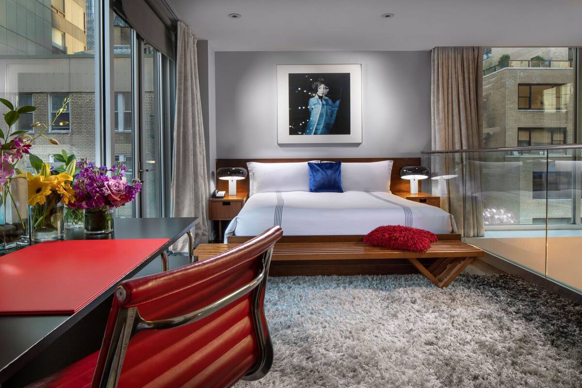 A modern hotel room with a large bed, a desk, and a chair. There is a window with a view of buildings outside.