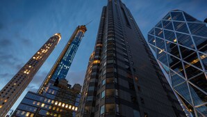 Um edifício alto com fachada de vidro moderna e design geométrico, cercado por outros edifícios imponentes.
