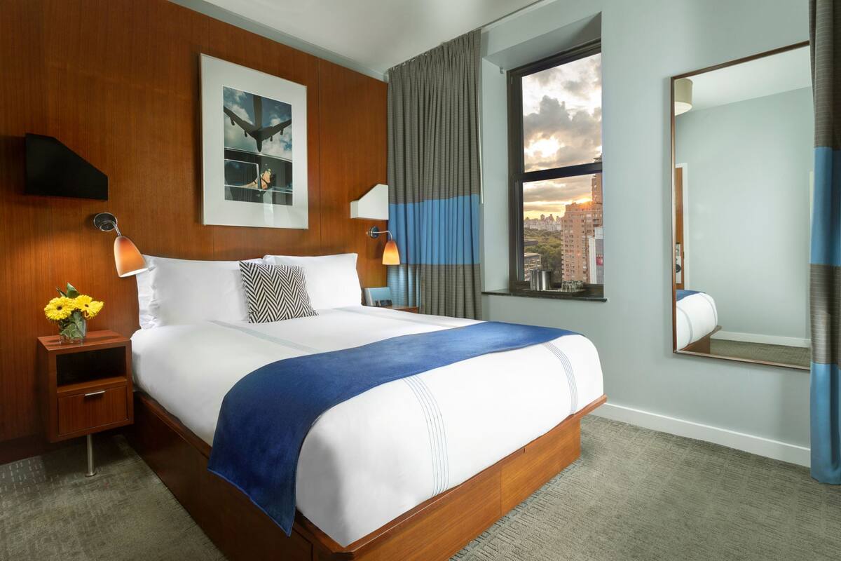 A hotel room with a large bed, a city view, a mirror, and a nightstand with a lamp and flowers.