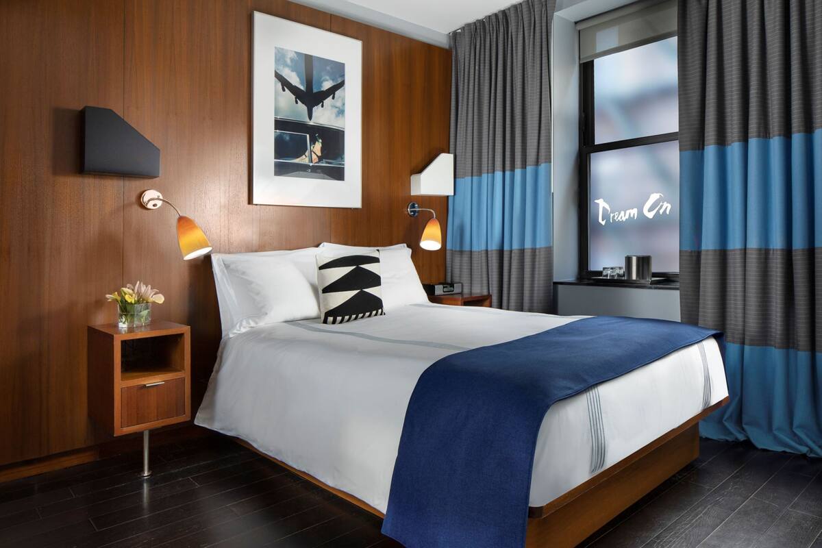 A modern hotel room with a large bed, wooden panel walls, a nightstand with a lamp, a small table with a lamp, and a window with curtains.