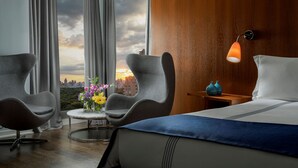 Quarto de hotel com cama, duas poltronas, uma mesinha com flores, mesa de cabeceira com abajur e vista para a cidade através de grandes janelas.