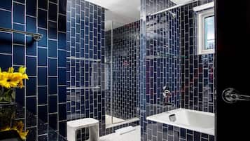 A modern bathroom with dark blue tiled walls, a glass shower enclosure, a white bathtub, and a small step stool.