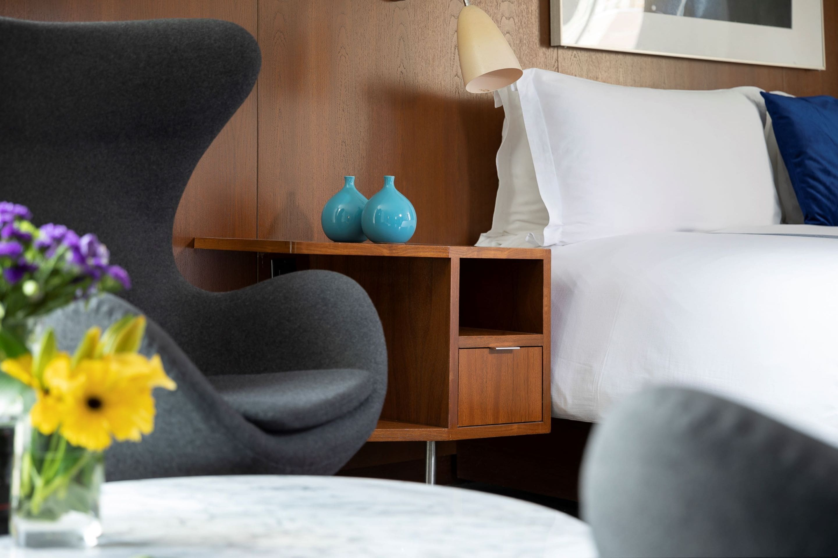 A modern hotel room with a wooden nightstand, a grey armchair, a white bed with a blue pillow, and a vase with yellow flowers.