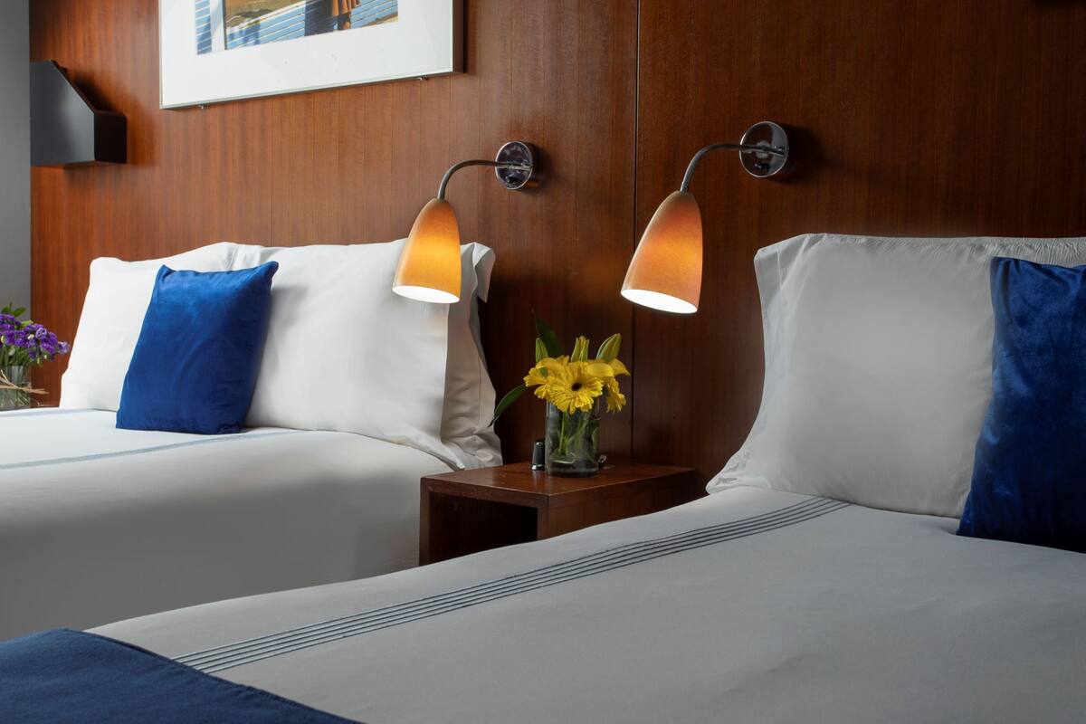 A hotel room with two beds, each with white linens and blue pillows, a small wooden nightstand with a vase of yellow flowers, and wall-mounted lamps.