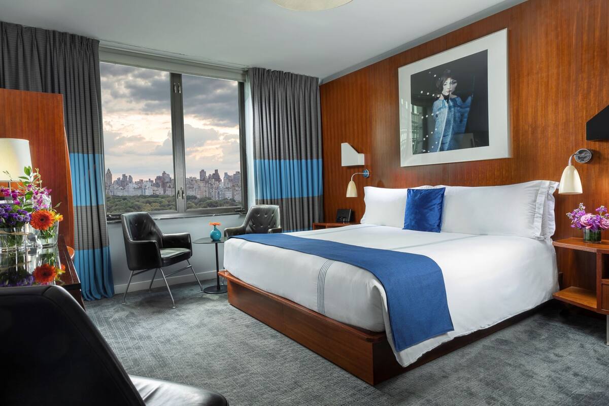 A hotel room with a large bed, a desk, a chair, a desk lamp, a nightstand, a framed picture on the wall, and a view of the city through the window.
