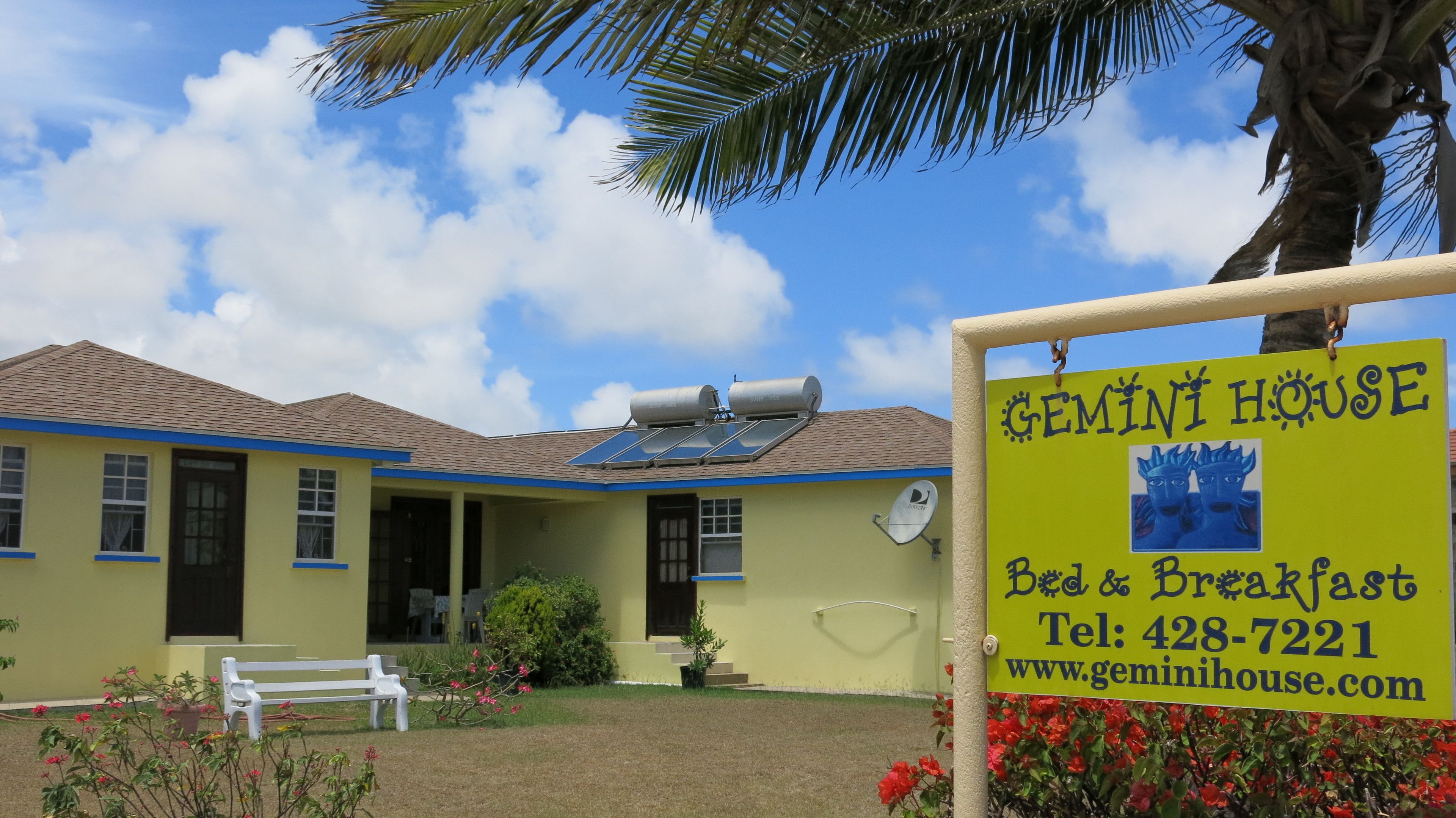 Ein gelbes Gebäude mit einem blauen Dach und Solarpanels. Ein Schild mit der Aufschrift: "GEMINI HOUSE Bed & Breakfast Tel: 428-7221 www.geminihouse.com".