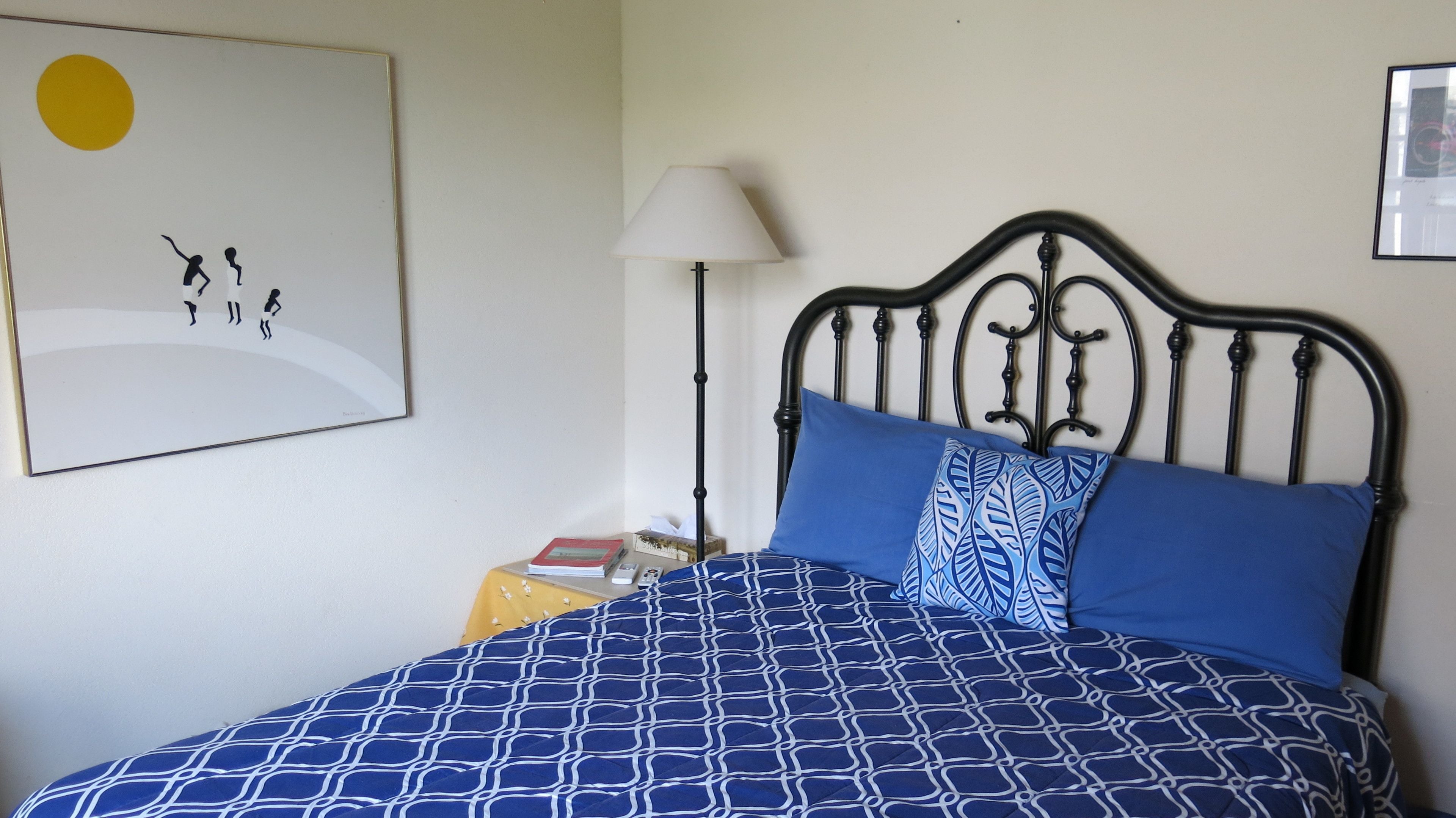A bed with a blue patterned bedspread and pillows, a bedside table with a lamp, and a framed picture on the wall.