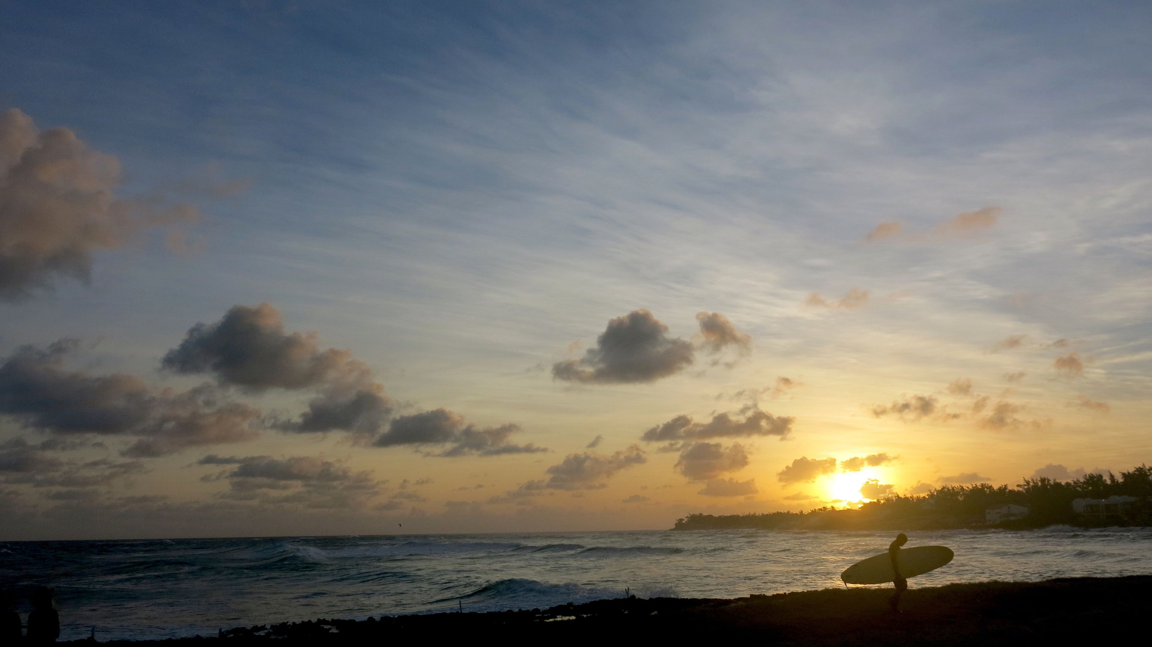 Cena de praia com um surfista carregando uma prancha, ondas do oceano e um pôr do sol ao fundo.