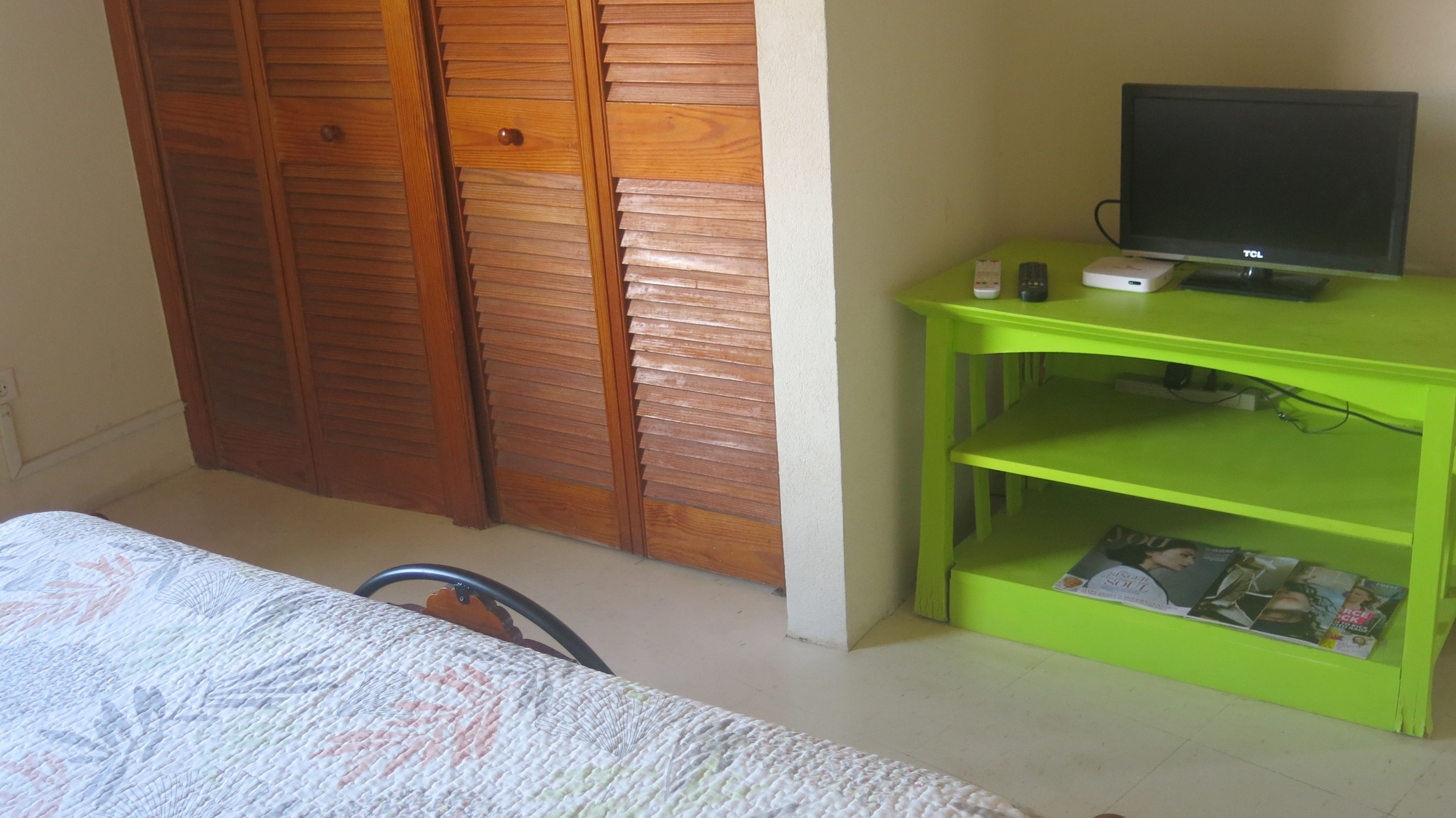 Ein Schlafzimmer mit einem Bett, einem Holzschrank, einer grünen TV-Konsole mit Fernseher und Fernbedienung.