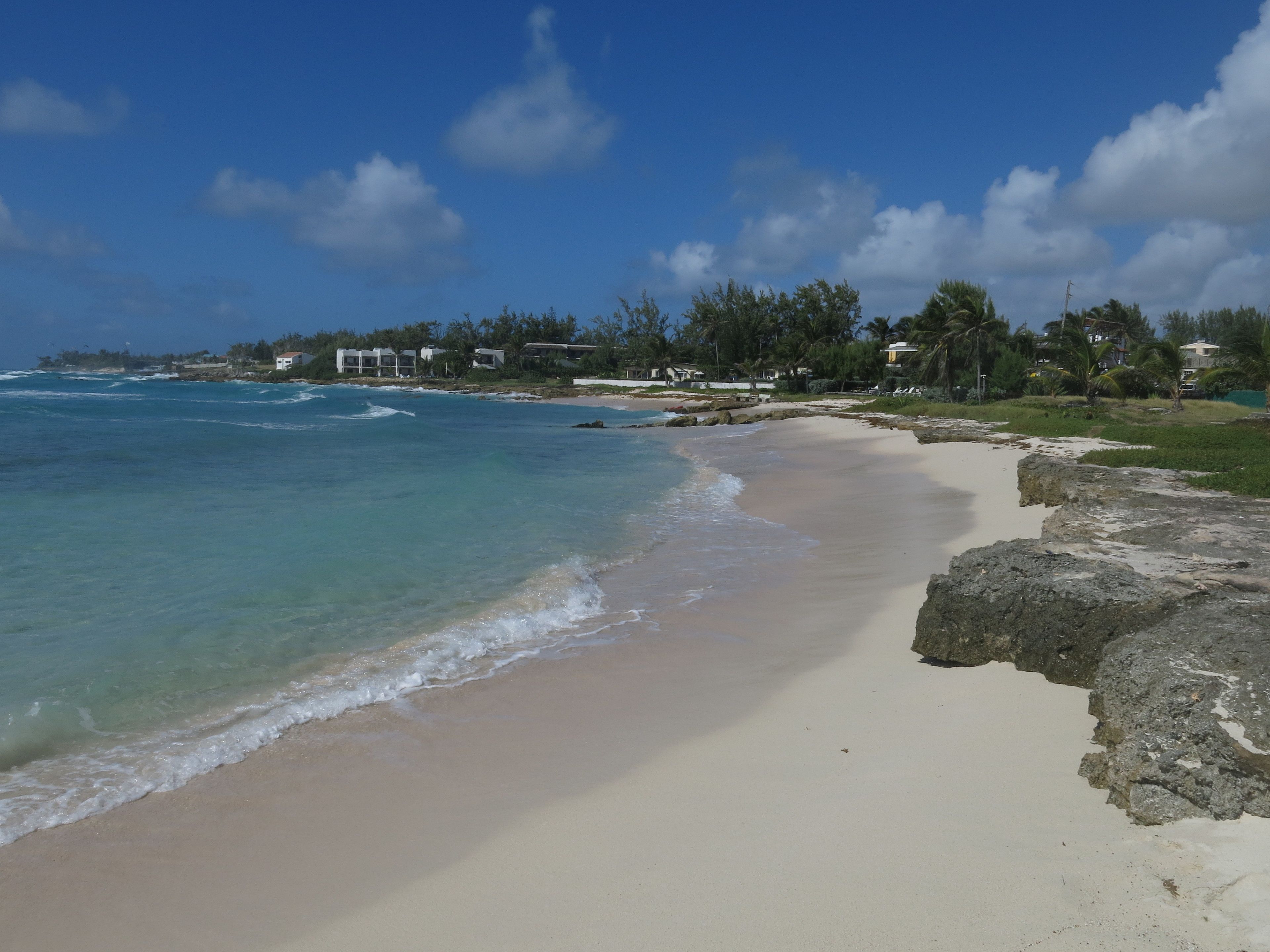 A beach with clear turquoise water, a sandy shore, and a few buildings in the background.