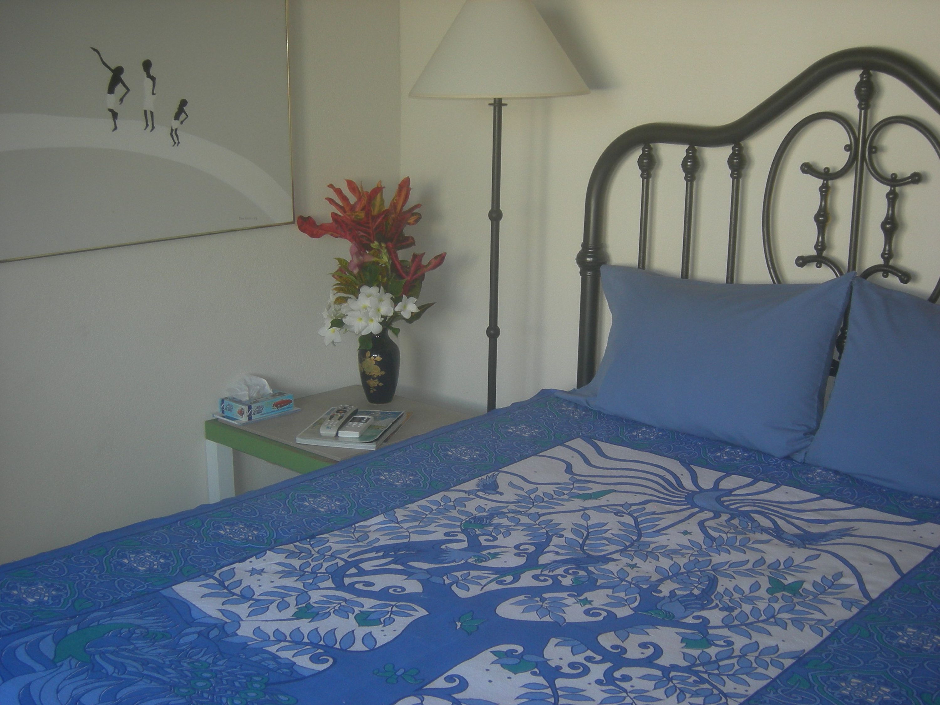 A neatly made bed with a blue and white patterned bedspread, a blue pillow, and a vase with flowers on a bedside table.