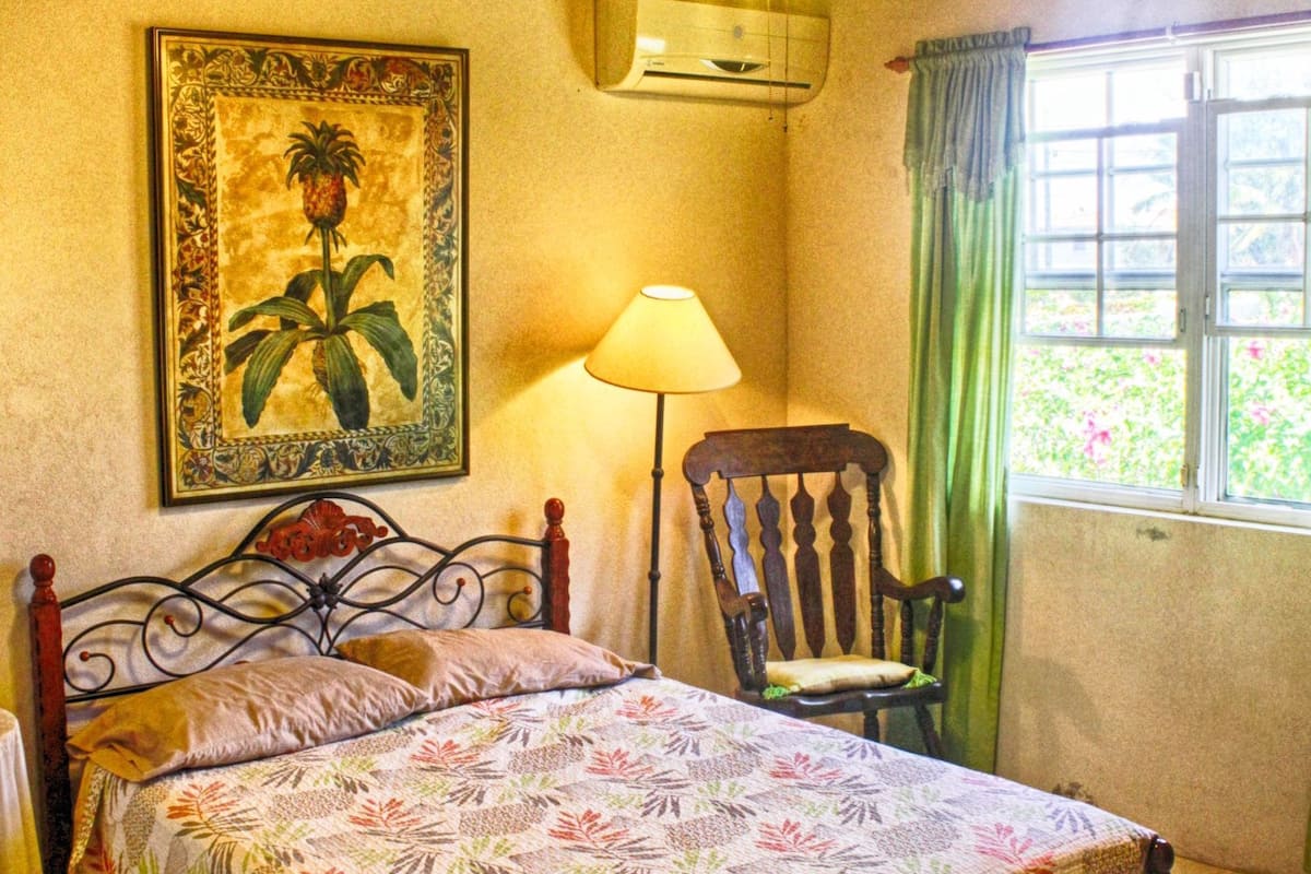 Quarto com teto de madeira, cama com colcha florida, uma cadeira, abajur e janela com cortinas.