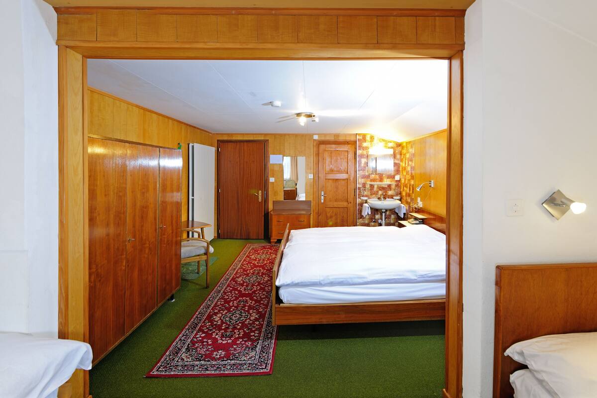 Ein Hotelzimmer mit einem Holzschrank, einem Bett mit weißen Leinentüchern, einem grünen Teppich und einem Badezimmer mit Waschbecken und Spiegel.