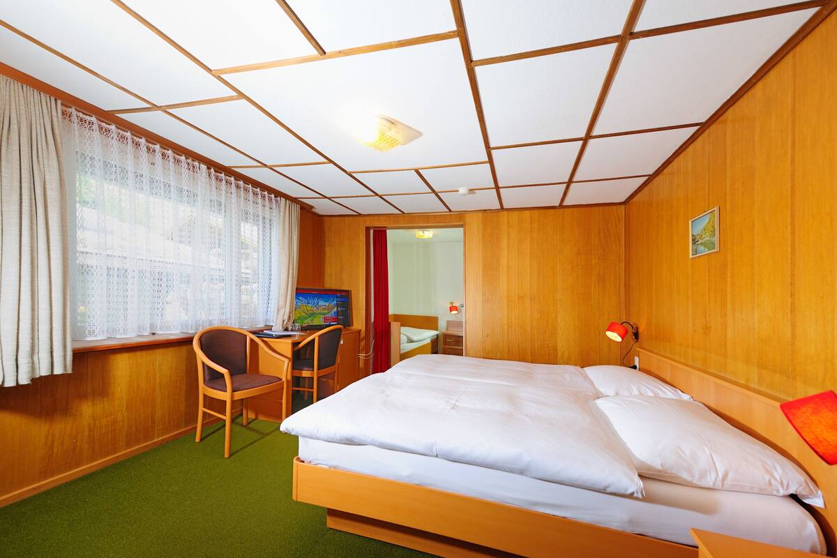 Ein Hotelzimmer mit einem Bett, einem Schreibtisch mit Stuhl, einem Computer und einem Fenster mit Vorhängen.