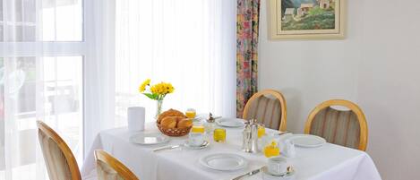 A dining table set for breakfast with a white tablecloth, plates, cups, and a basket of croissants.