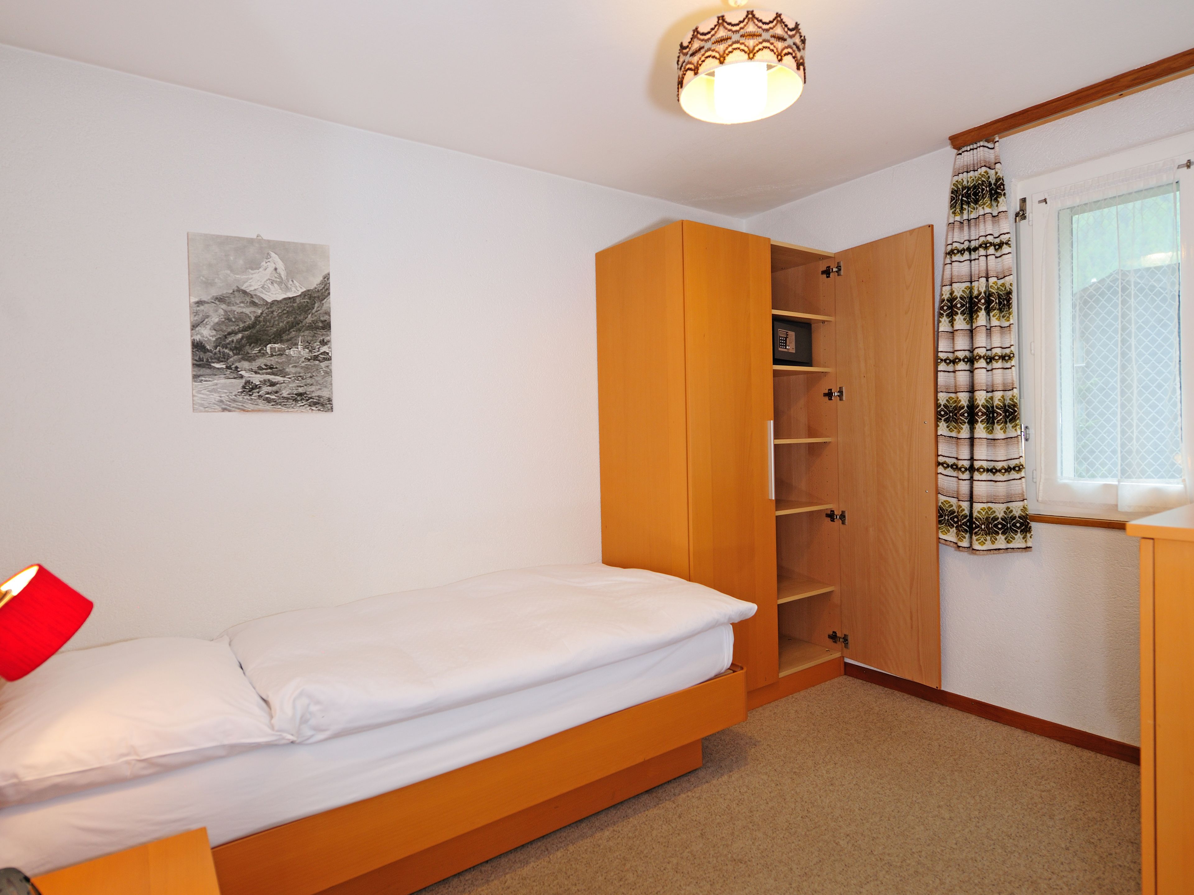 Un dormitorio con una cama individual, un armario, una estantería, un cuadro colgado en la pared y una ventana con cortinas.