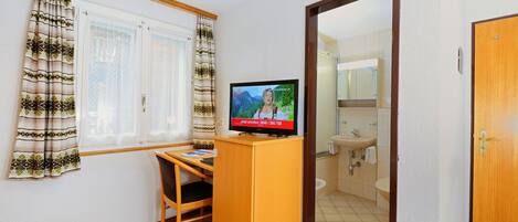 Ein kleines Zimmer mit einem Holztisch, einem Fernseher, einem Fenster mit Vorhängen und einem durchgegangenen Türspalte sichtbaren Badezimmer.