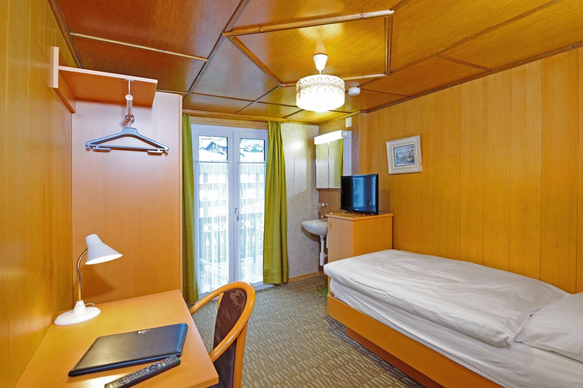 Ein Hotelzimmer mit Bett, Schreibtisch mit Laptop und Fernbedienung, Fernseher, Kleiderhaken und einer Dusche.