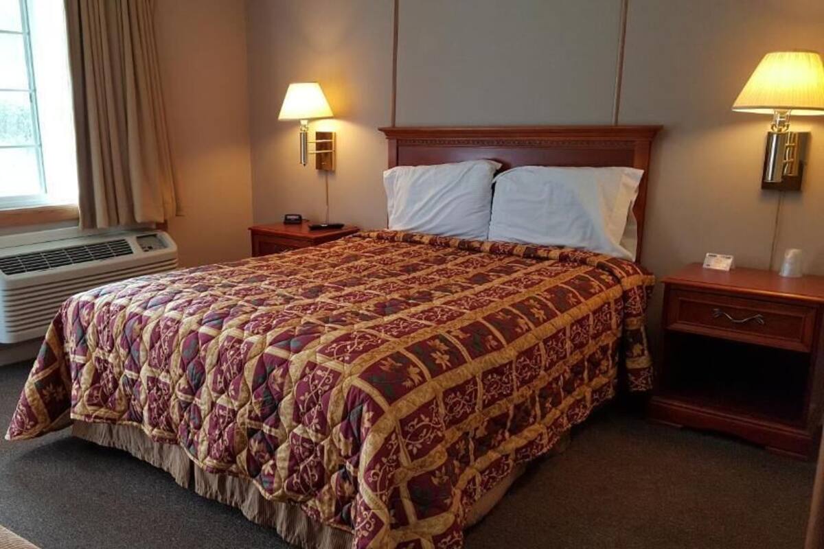 A hotel room with a large bed, two bedside tables, a window with curtains, an air conditioning unit, and two wall-mounted lamps.