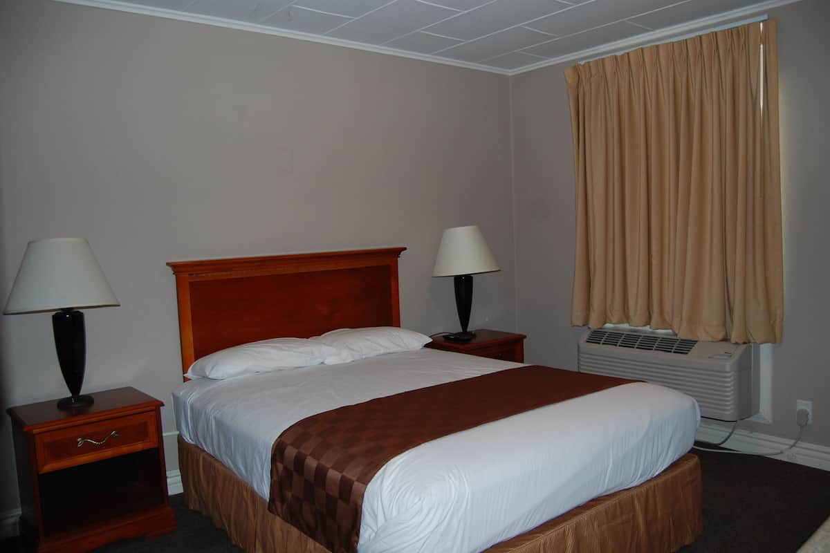 A hotel room with a single bed, two bedside tables with lamps, a window with curtains, and an air conditioning unit.