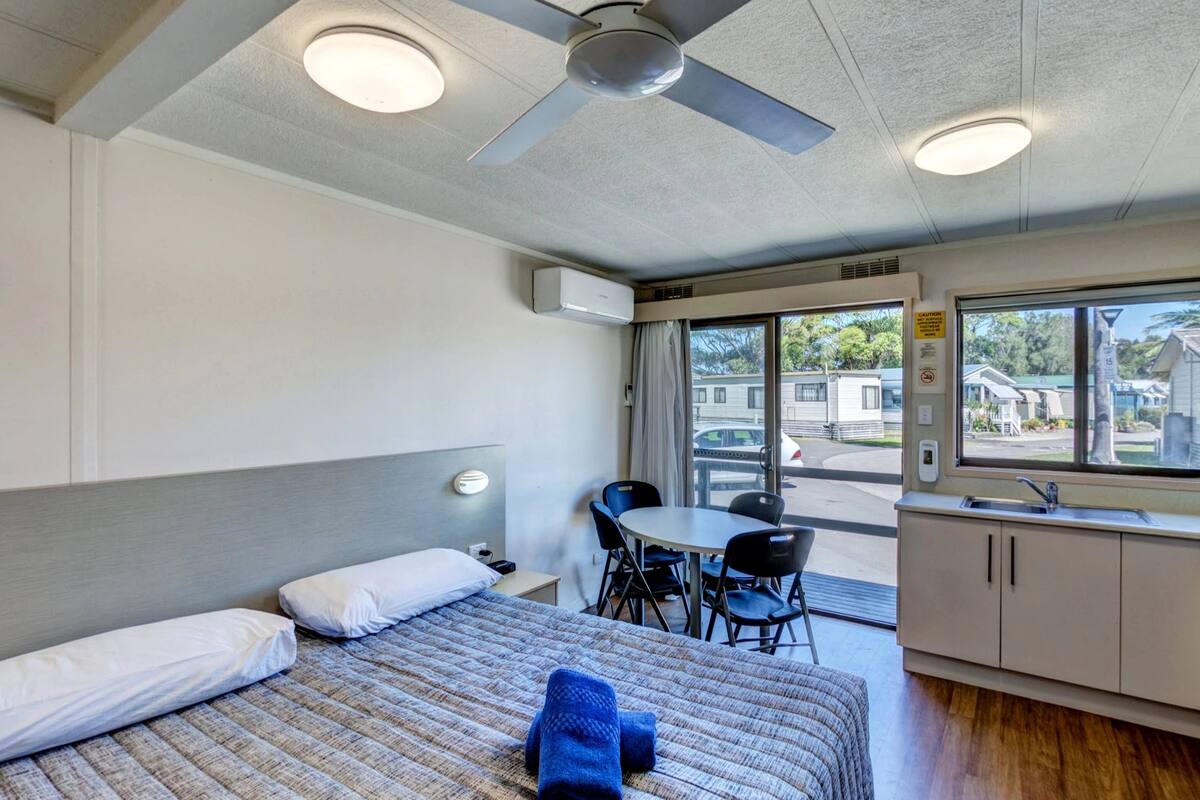 A hotel room with a bed, a desk, a chair, a kitchenette, and a window with a view of the outside.